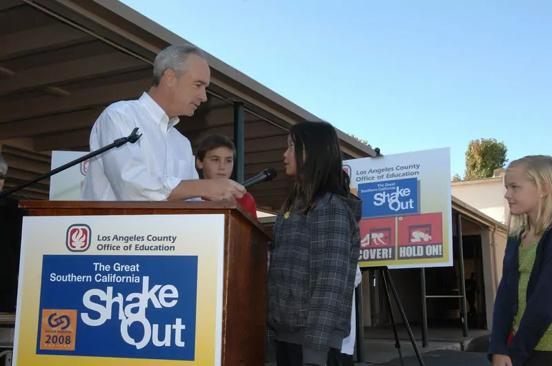 A man at a podium hands a microphone to a schoolgirl in a grey jacket, in front of signs for the Great Southern California ShakeOut 2008