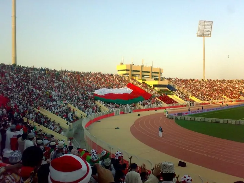 Thousands of Omani fans in red and white fill a stadium stand in Muscat, with a giant Oman flag spread across the seats and a running track beside the pitch