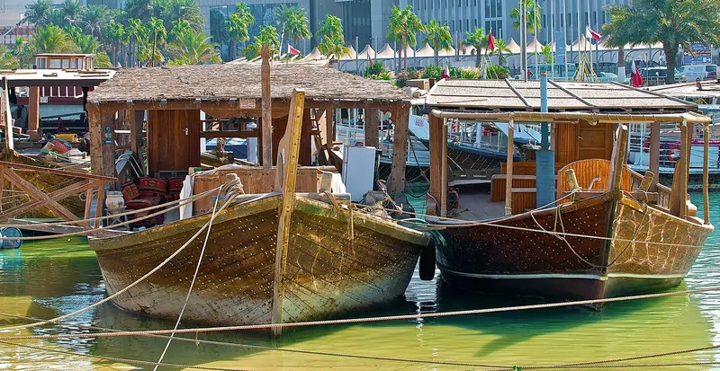 Two traditional wooden dhows with thatched shade roofs moored in green water, with palm trees behind