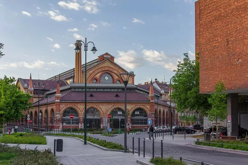 The red brick back facade of the Great Market Hall in Budapest with a green and gold tiled roof, seen from a quiet street