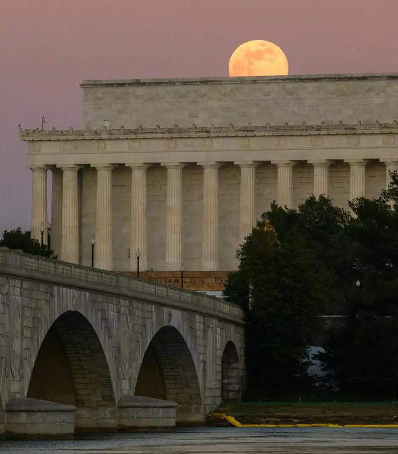 An orange full moon rising just above the roof of the Lincoln Memorial, with the Memorial Bridge arches in front