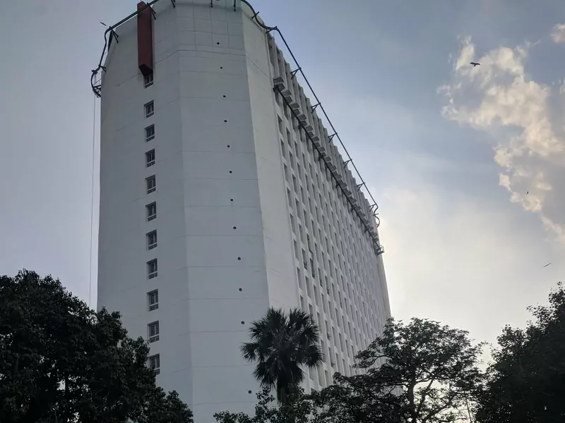The tall white tower of Sanchar Bhawan in New Delhi, seen from the road through trees and a palm, under a hazy sky with birds