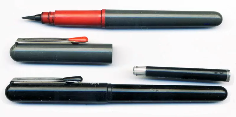 Two black brush pens, one uncapped with a pointed felt tip and a red grip, laid out on a white background with their caps and a spare ink cartridge
