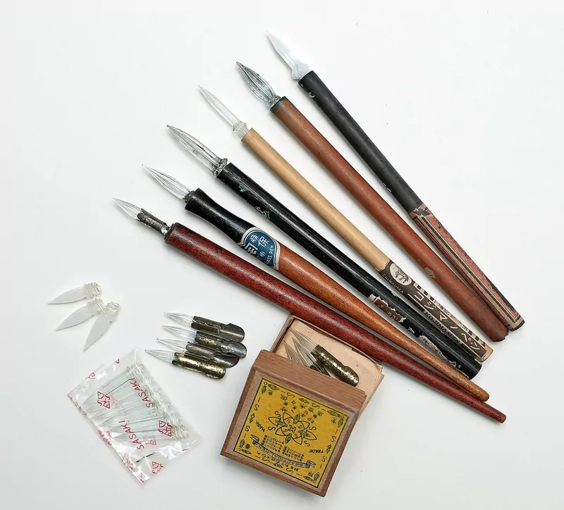 Several wooden dip pen holders with glass nibs, spare metal nibs and a small box laid out on a white table