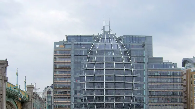 A tall glass and steel office building on the River Thames in London with an egg-shaped glass frame, beside a stone bridge under a cloudy sky