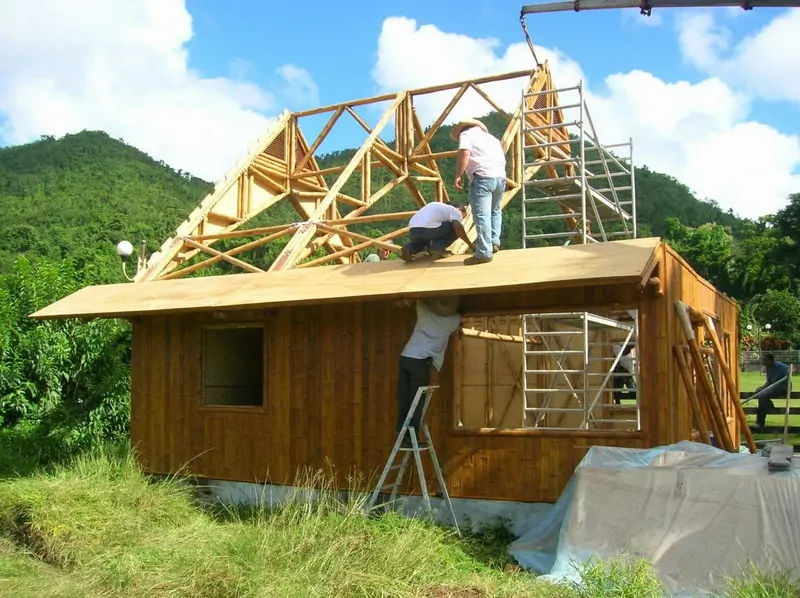 Workers fit the timber roof frame of a small wooden house on a green hillside