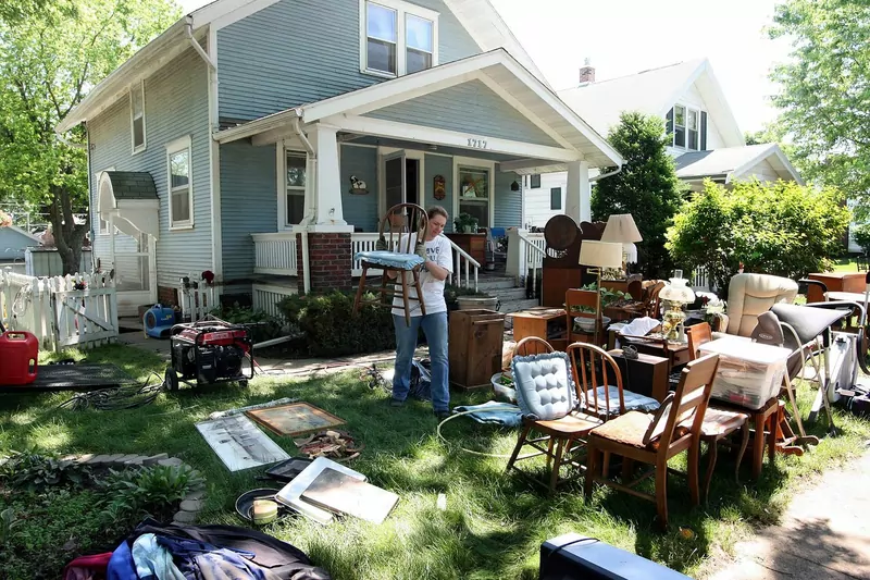 A person stands on the front lawn of a house among armchairs, a table, lamps and other furniture laid out after a flood