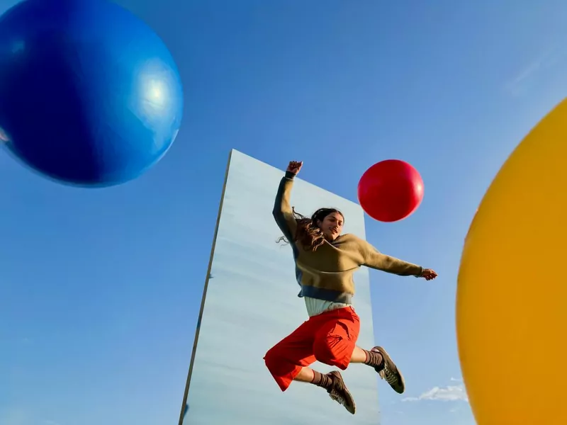 A man in red trousers leaping past large blue, red and yellow balloons against a blue sky