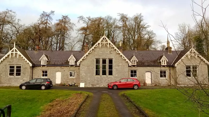 A long grey stone school building with three pointed gables and two cars parked in front, behind a green lawn and autumn trees