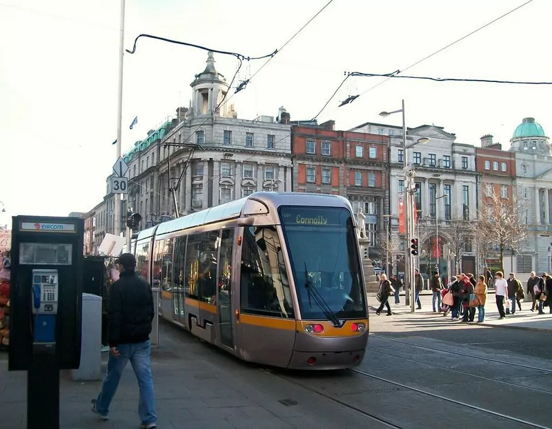 A silver and yellow Luas tram bound for Connolly at the junction of Abbey Street and O'Connell Street in Dublin, with people walking past