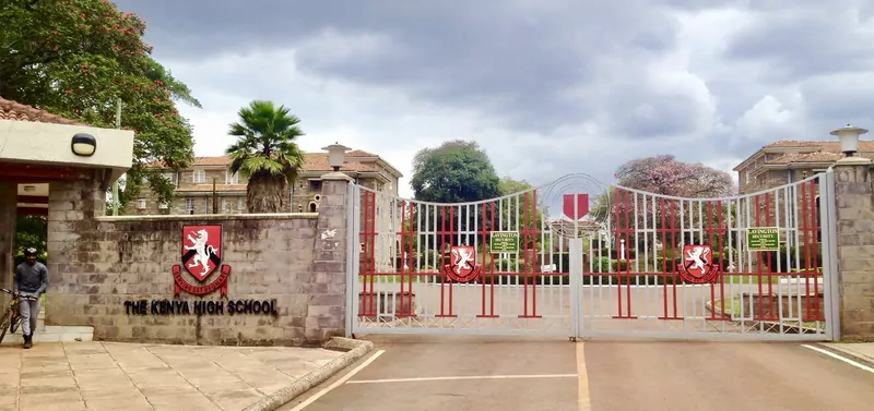 The red and white gates and brick gatehouse of the Kenya High School in Nairobi, with the school crest on the wall and a man walking his bicycle at the left