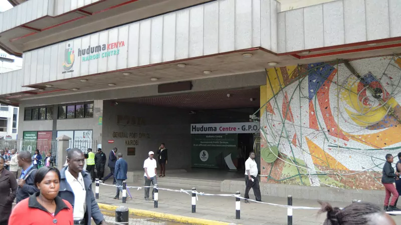 The entrance of the Huduma Centre at the General Post Office in Nairobi, with the Huduma Kenya sign over the door, a colourful mosaic wall and passers-by in coats and jackets on the pavement