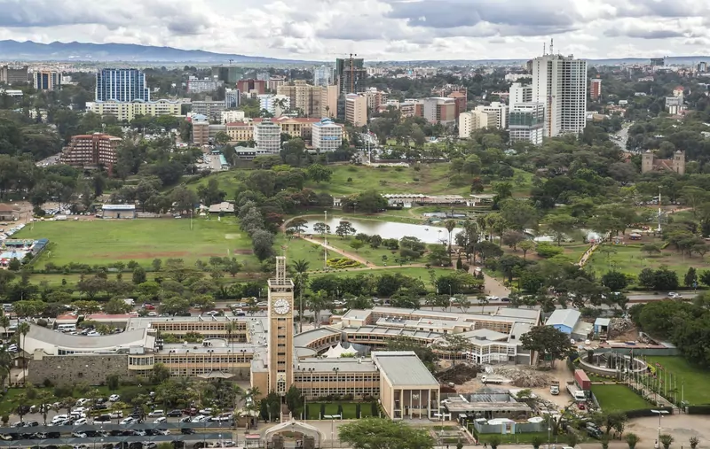 The clock tower and low wings of the Parliament Buildings in Nairobi in the foreground, with the green lawns and lake of Uhuru Park and the city centre behind