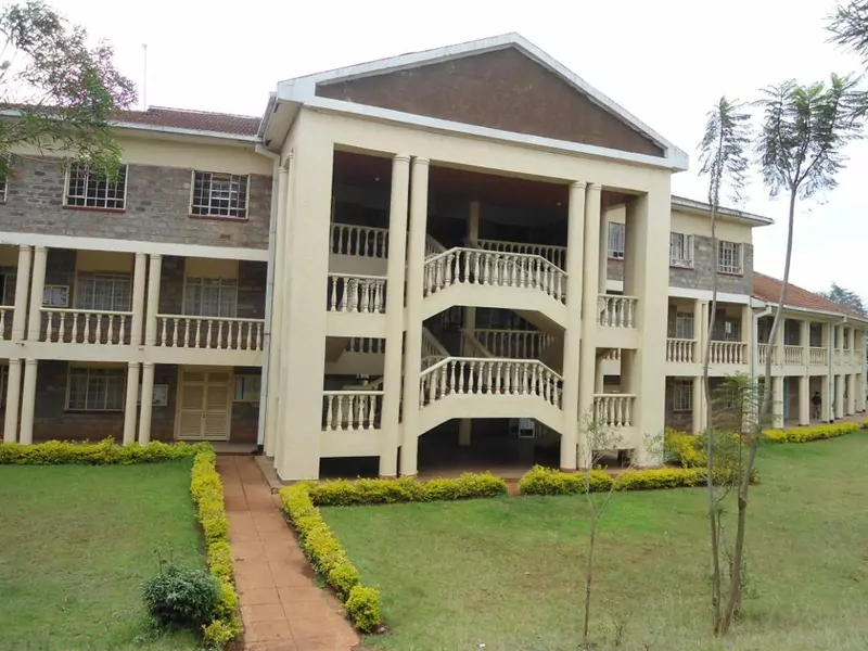 A cream-coloured three-storey school administration block with white balustraded balconies, a brick-clad side wing and a pitched entrance gable, with trimmed hedges and a paved path in front