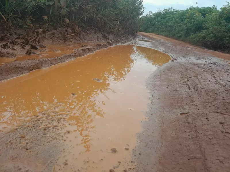 A dirt road in Siaya County with a long puddle of orange flood water across it, the reflections of leaves on the surface, red mud at the edges and green bush on both sides
