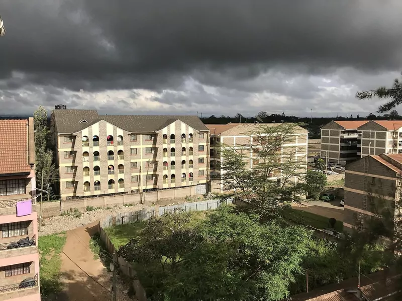 Dark grey storm clouds hang low over blocks of flats with cream and brown walls, trees and a dirt path in Nairobi, with sunlight on the buildings