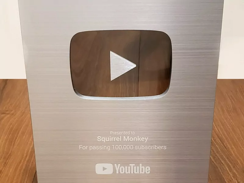 A brushed-silver YouTube award plaque on a wooden table, with a play-button cut-out, the words &ldquo;For passing 100,000 subscribers&rdquo; and the YouTube logo