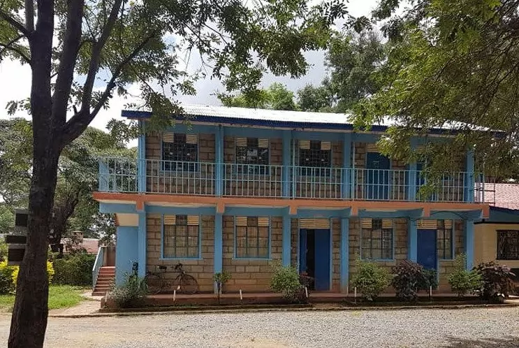 A two-storey school library and IT building with blue pillars, brick walls and a balcony, with a bicycle parked by the entrance, in Mwala, Kenya