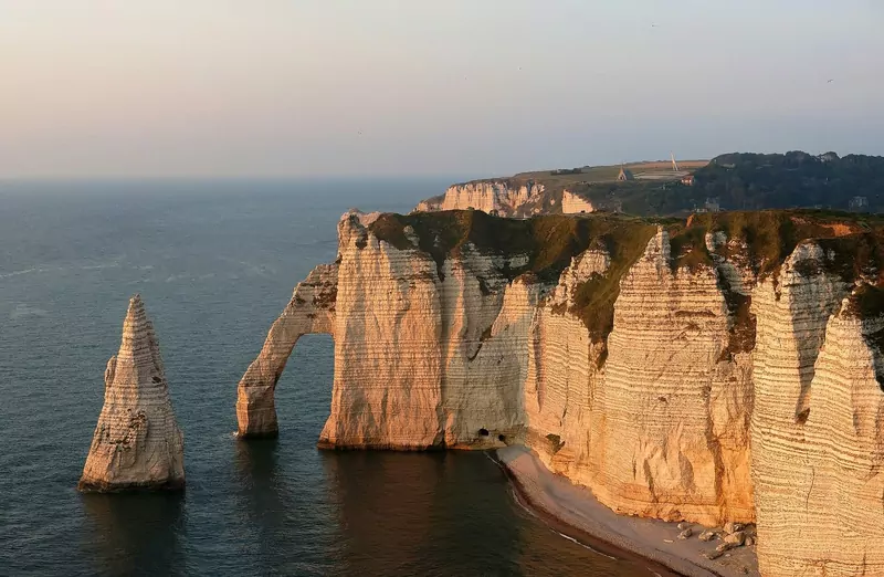 White chalk cliffs with a natural rock arch and a tall needle-shaped rock standing in the sea, lit orange by the evening sun