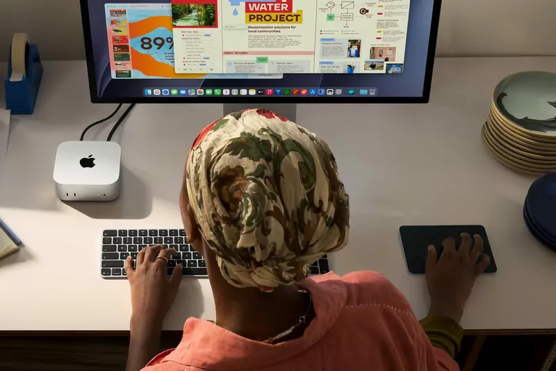 A person in a patterned head wrap and coral shirt typing at a desk with a Mac mini, a monitor and a trackpad