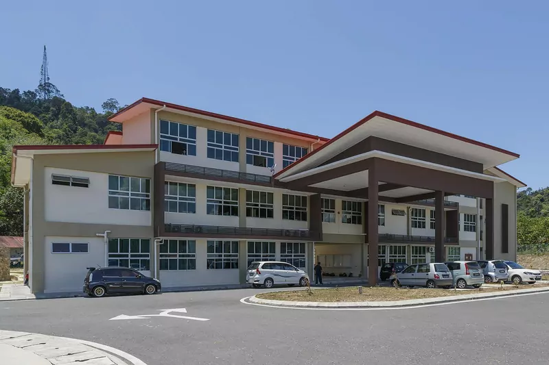 A modern three-storey primary school building in Sandakan, Sabah, with cream walls, brown pillars, a wide covered entrance, parked cars and forested hills behind