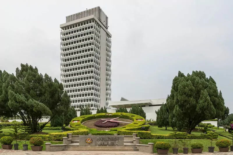 The Parliament of Malaysia tower in Kuala Lumpur behind a clipped hedge flower clock and a sign reading Parlimen Malaysia