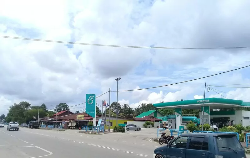 A Petronas station beside a road in Pagoh, Johor, with its teal canopy and sign under a cloudy sky, a small dark blue car in front and a motorcyclist behind it
