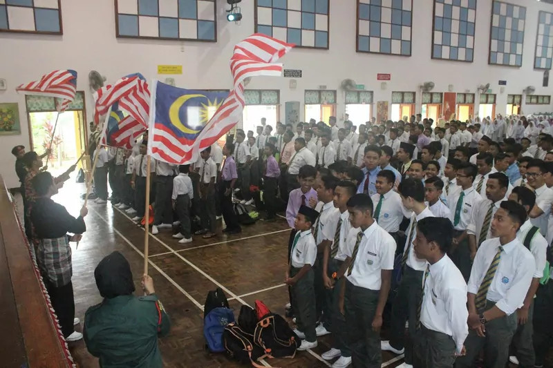 Rows of boys in white shirts, ties and grey trousers stand in a school hall while students at the left wave large Malaysian flags