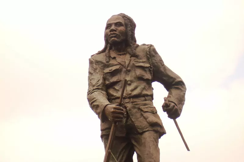 A bronze statue of Dedan Kimathi in a military jacket with long dreadlocks, holding a rifle and a dagger, seen from below against a pale sky