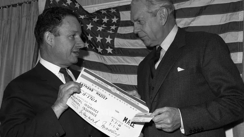 Black-and-white photo of two men in suits in front of a US flag, one handing the other a large Medicare identification card