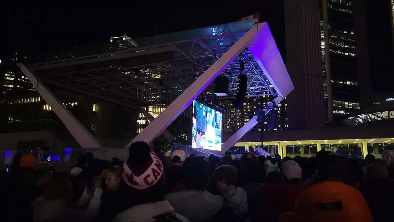 A large crowd at night in Nathan Phillips Square in Toronto, watching a baseball game on a big outdoor screen