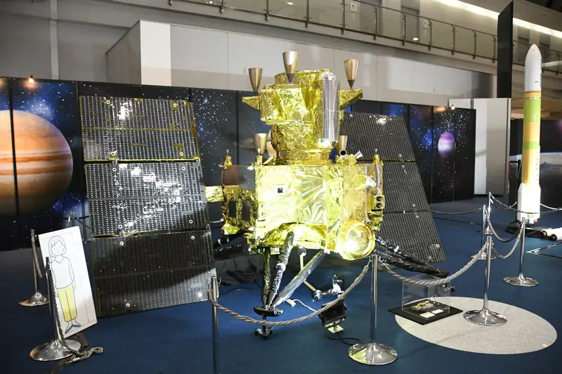 A half-scale model of the MMX spacecraft with gold foil and solar panels on display at an exhibition