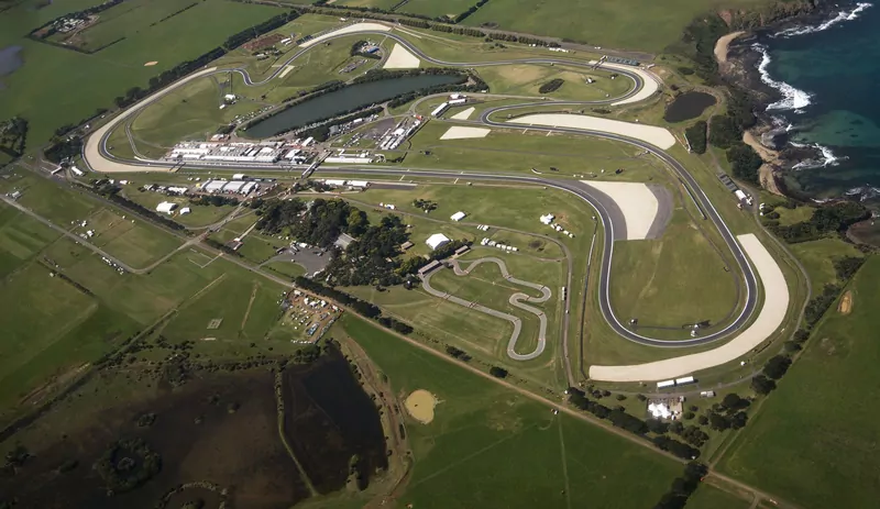 Aerial view of the Phillip Island Grand Prix Circuit in Victoria, Australia: a looping ribbon of track around a lake and pit buildings, with green farmland and a rocky, wave-washed coast on the right
