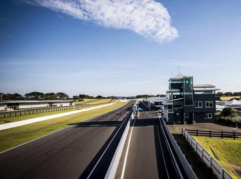 The empty main straight and pit lane at Phillip Island, with a dark race-control tower on the right, a long grassy run-off on the left and a clear blue sky with one streaky cloud