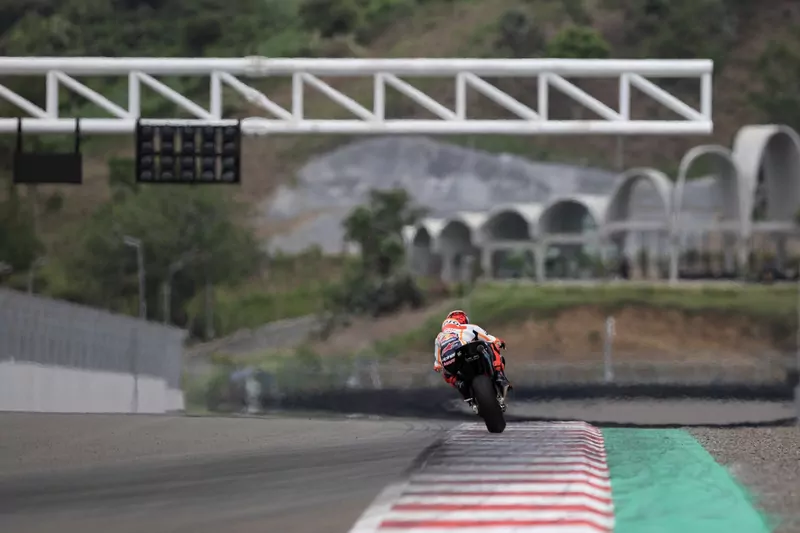 A MotoGP rider in orange and white Repsol Honda leathers accelerates away down the main straight at Mandalika, with the circuit's white arched buildings on the hillside behind