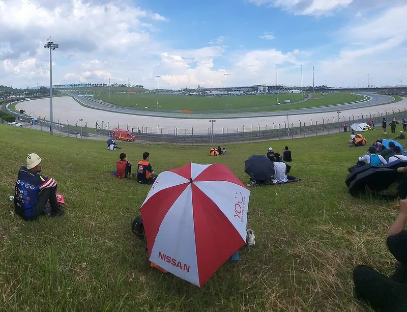 Spectators sit on a grassy bank under palm trees at the Sepang International Circuit, with a red and white umbrella in the foreground, a wide curve of the track and run-off area below and a blue sky with large clouds