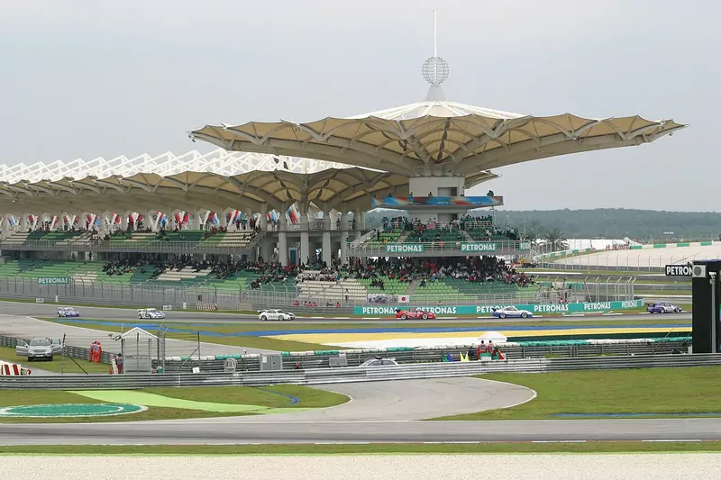 The curved, sail-like roof of the main grandstand at the Sepang circuit above green seating and Petronas banners, with racing cars on the track in front