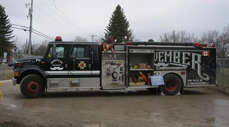 A black fire truck painted with a large Movember logo and moustache designs, parked on a gravel lot