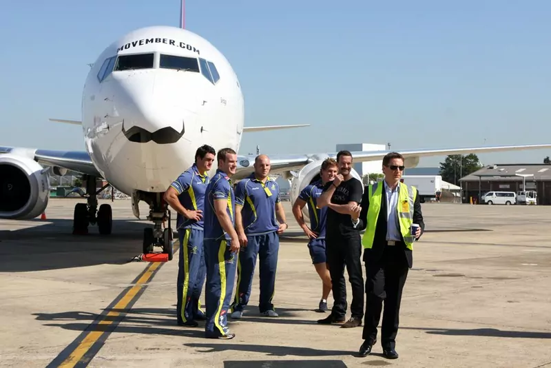 A white passenger jet with a giant black moustache painted under its nose, with rugby players standing in front of it