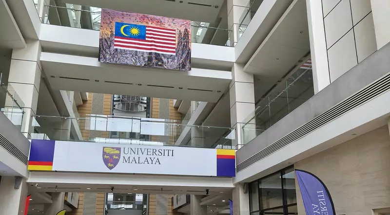 A tall glass-roofed lobby with a large Malaysian flag banner hanging above a white Universiti Malaya sign and purple and yellow banners
