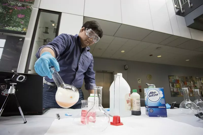 A demonstrator in safety goggles pours a liquid from a round flask at a table full of bottles and beakers