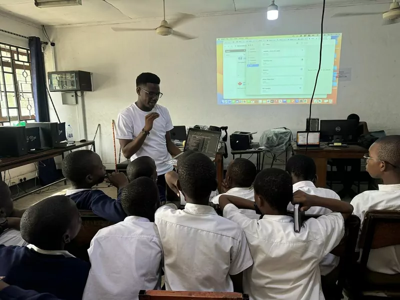 A teacher presenting on a projector screen to students in white shirts in a computer room
