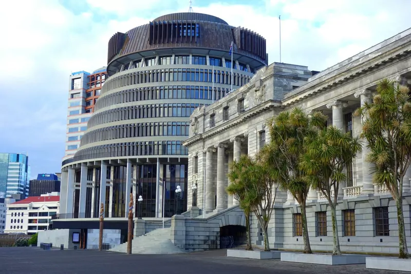 The round Beehive building of the New Zealand Parliament next to the stone Parliament House in Wellington, with cabbage trees in front and carved wooden posts at the entrance