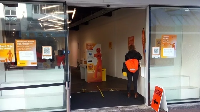 The open glass door of an advance voting place in Wellington with orange Electoral Commission posters reading Enrol. Vote. Be heard., a voting sign on the pavement and a voter in a dark jacket and orange backpack at the desk