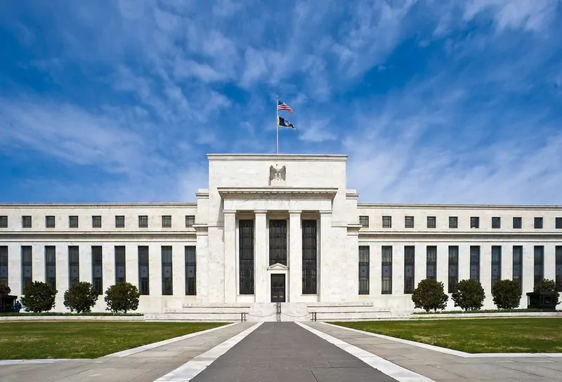 The white marble front of the Federal Reserve's Eccles Building under a blue sky, with a flag on top and lawns in front