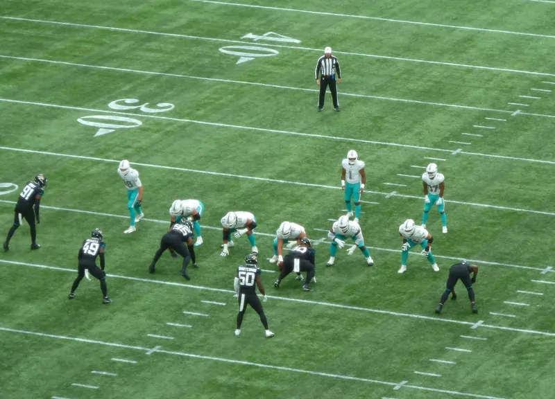 Jaguars players in black and Dolphins players in white line up on the green field at Tottenham Hotspur Stadium, with a referee behind them