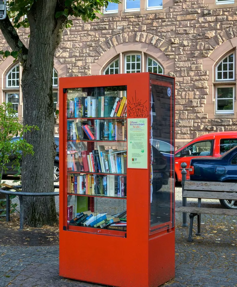 A red public bookcase with glass doors full of books, standing on a cobbled square beside a tree trunk and a wooden bench, in front of a stone building
