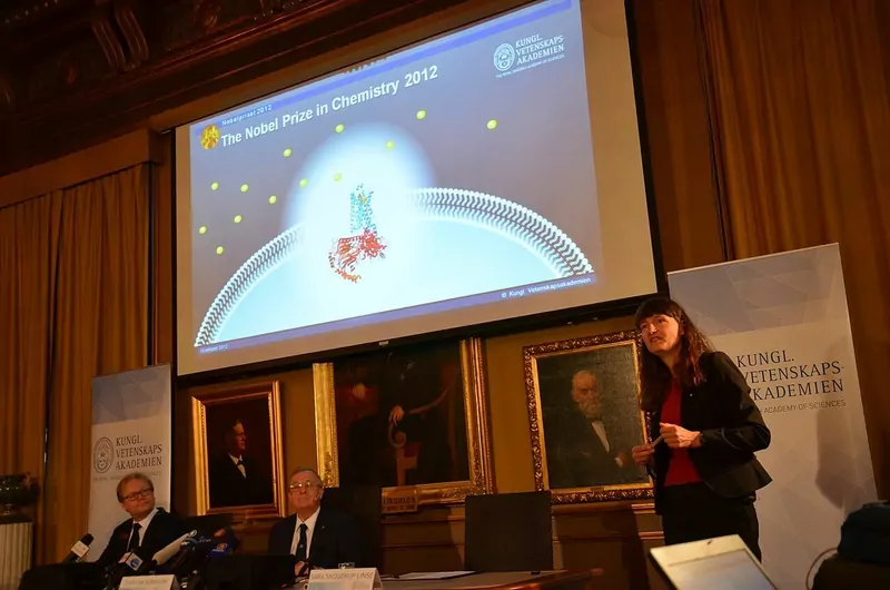 A scientist presents the chemistry prize research in front of a large screen at a wood-panelled hall, with two committee members seated at a table
