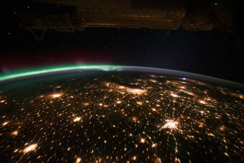 View from the International Space Station of a green aurora glowing along Earth's horizon above the city lights of the American Midwest at night