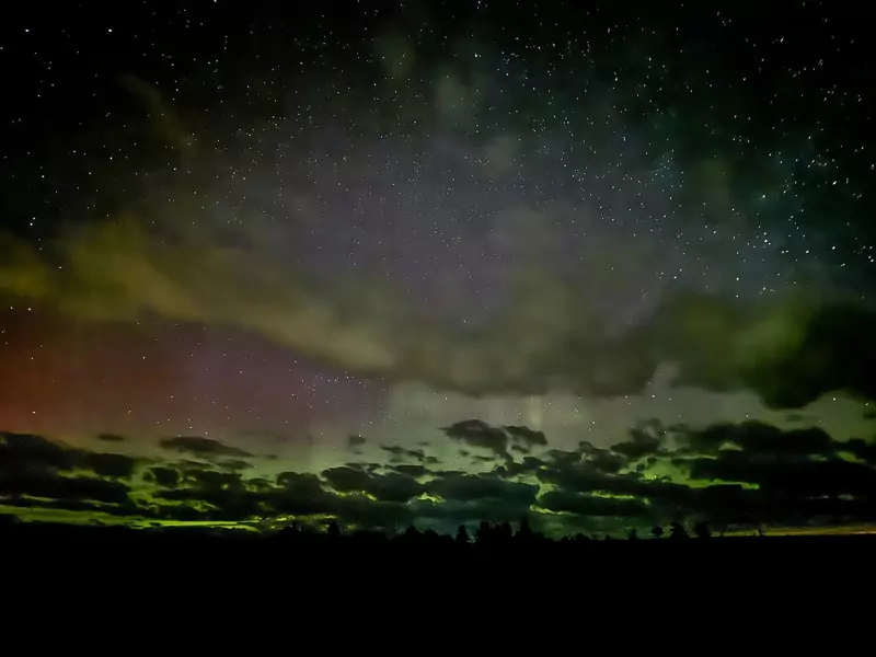 Faint green and red glow of the northern lights low over a dark treeline and field in Maine, with stars above